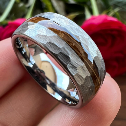 A hammered metal ring held on a person's fingers, with blurred red roses in the background. The ring has a polished silver-toned band with a textured, hammered finish and a central brown inlay.